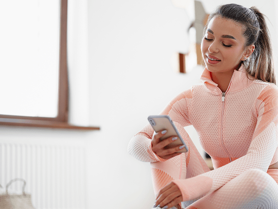 Young woman in sportswear sitting indoors and looking at smartphone before workout